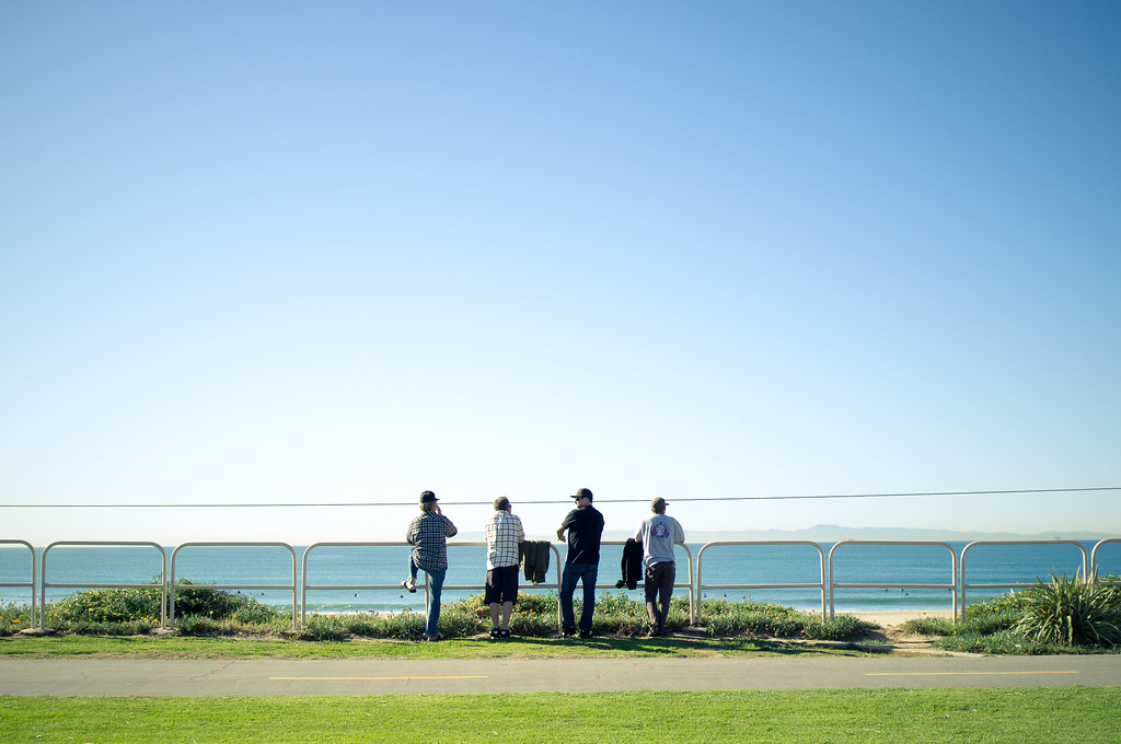 Four men leaning against a railing overlooking the calm Pacific Ocean from a grassy California park.