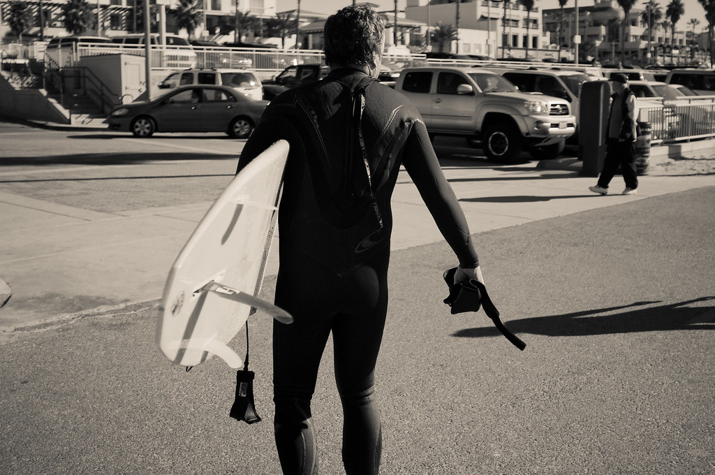 Black and white view of a surfer in a wetsuit carrying a surfboard along a coastal California street.