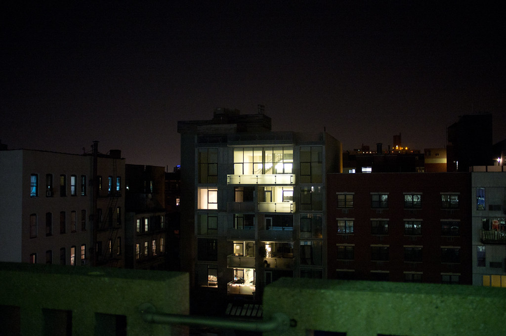 Nighttime rooftop view of an illuminated glass apartment building against a dark city sky in New York.