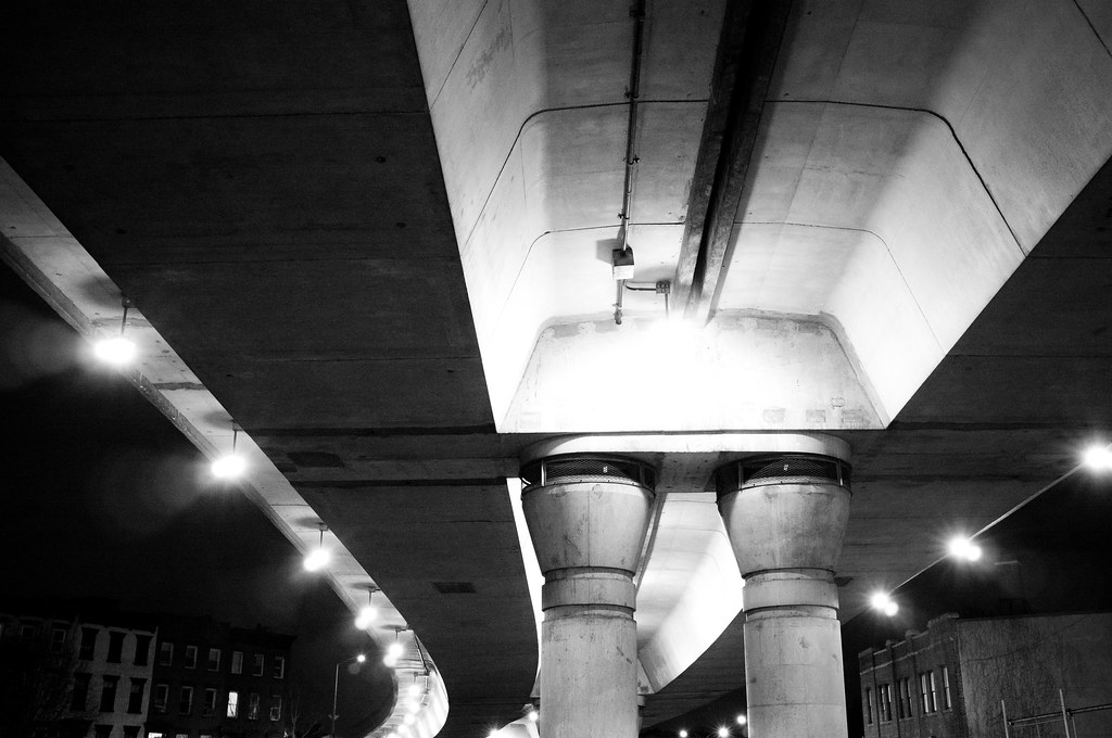 Illuminated concrete pillars and curved ramps of an elevated highway overpass viewed from below at night.