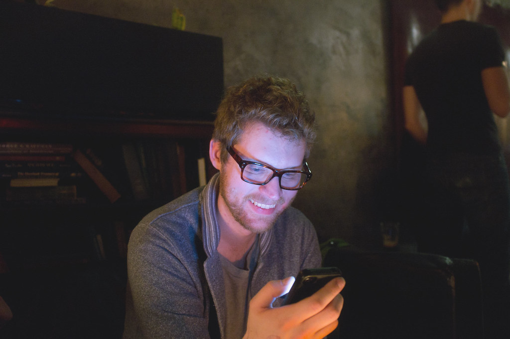 Ren Bostelaar smiling while looking down at his glowing smartphone screen in a dimly lit bar.