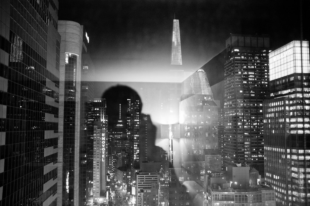 High-rise window view of illuminated Midtown Manhattan skyscrapers at night with a reflected silhouette.