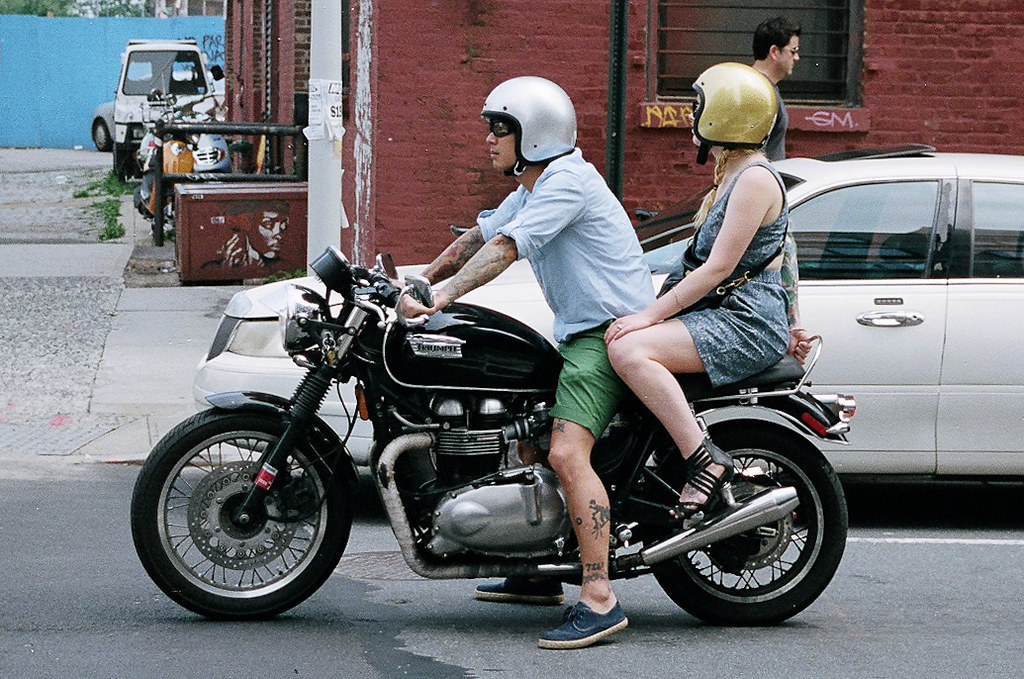 A man and woman wearing silver and gold motorcycle helmets riding a black Triumph motorcycle on a Brooklyn street.