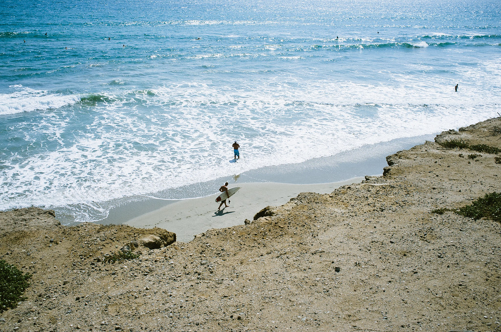 High-angle view from a dirt cliff overlooking surfers in the ocean along the sandy California shoreline.