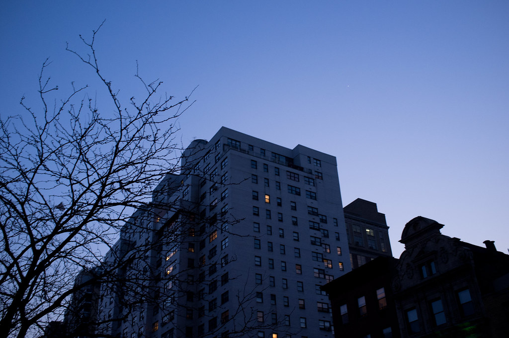 Apartment building with lit windows at dusk framed by silhouetted tree branches against an evening sky.