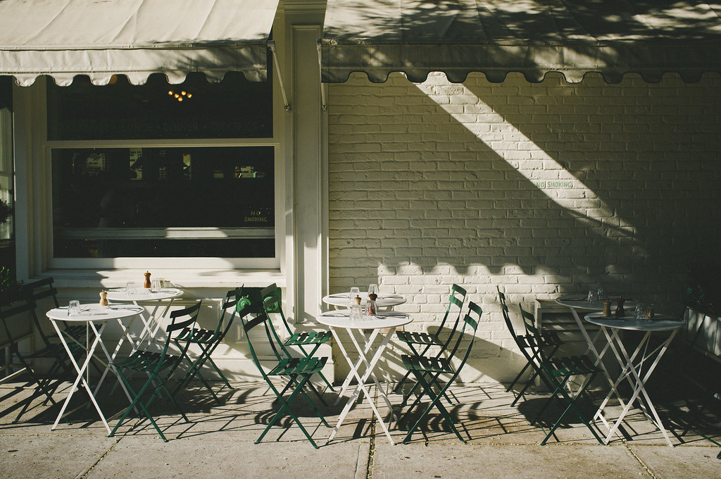 Sidewalk dining tables with green folding bistro chairs beneath an awning by a white brick cafe wall.