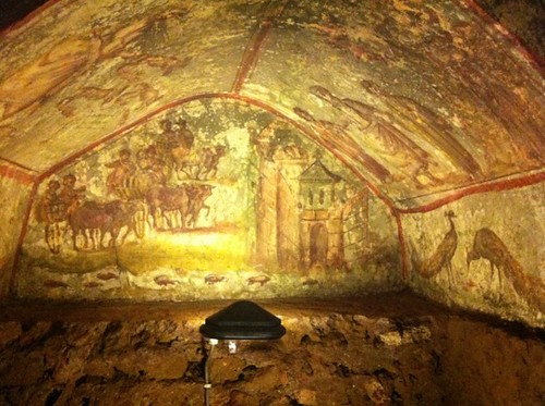 Catacombs in Via Latina, Rome: Underground tomb with a light in the center. The light illuminates the ceiling, which is covered in paintings of horses, men, and buildings. 