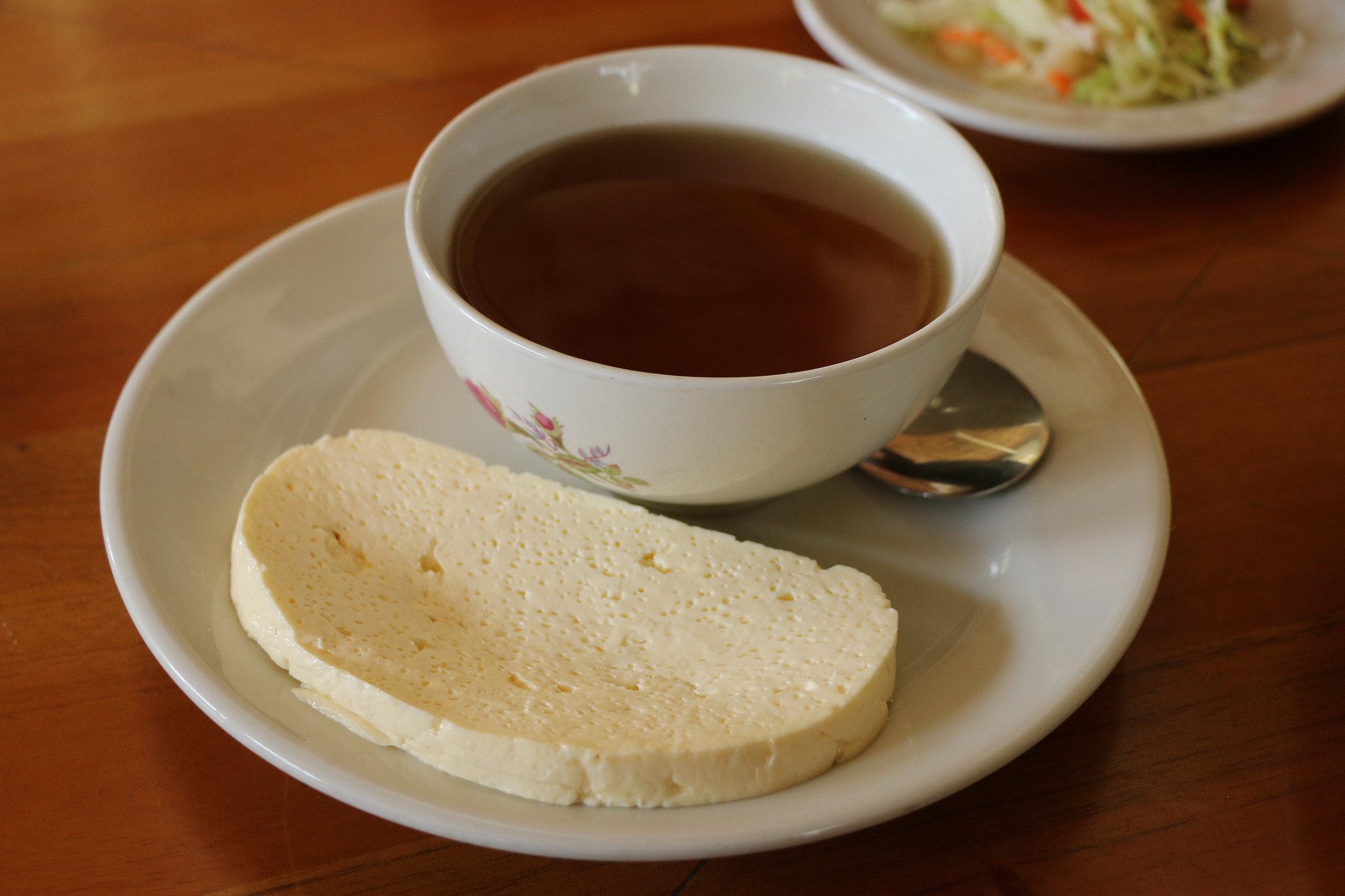 Plato pequeño blanco donde posa una cuchara y una tajada de queso y encima del plato un pocillo pequeño con panela infundida en agua como parte de los Platos Típicos del Eje Cafetero