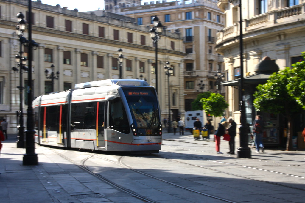 A multi-car tram runs on rails as it turns from a city street into a plaza