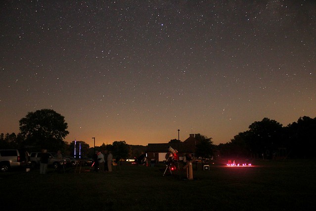 Starry sky with sunset still happening under it and a line of trees under it, people with telescopes and red lights are vaguely seen at the bottom