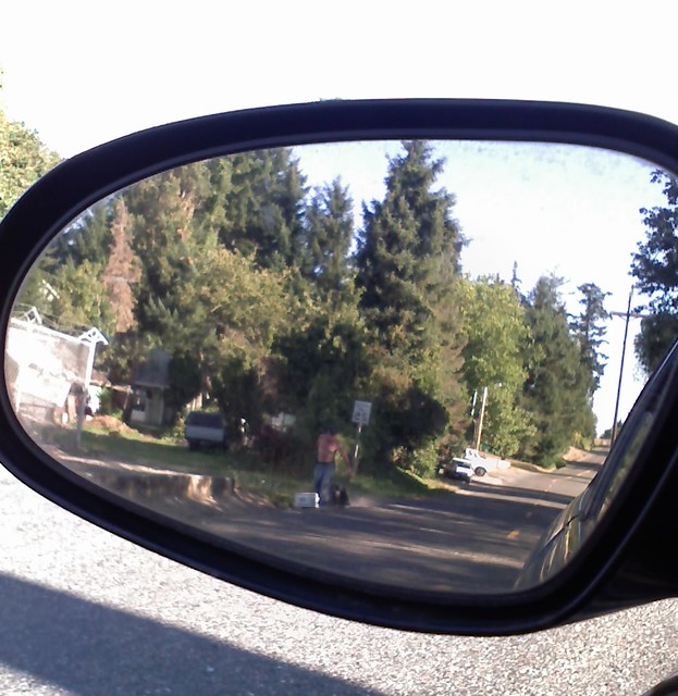 a man dragging a television and a microwave by their cords down the hill on the gravel, as seen through my rearview mirror