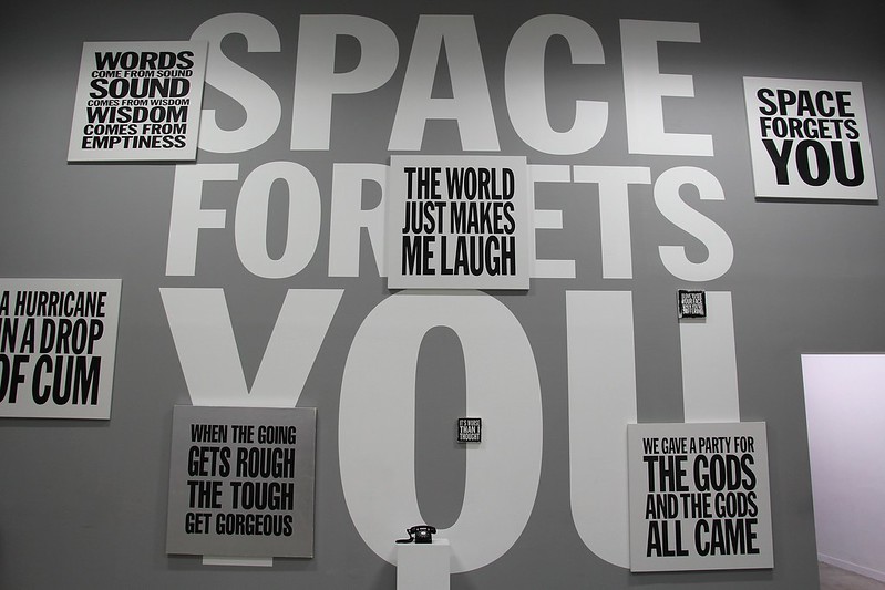 A photo of an art exhibit of John Giormo poetry paintings. The wall has large block text on it that says "Space Forgets You" and then small square paintings are over it saying things like "the world just makes me laugh", "we gave a party for the gods and the gods all came" and "when the going gets rough the tough get gorgeous"