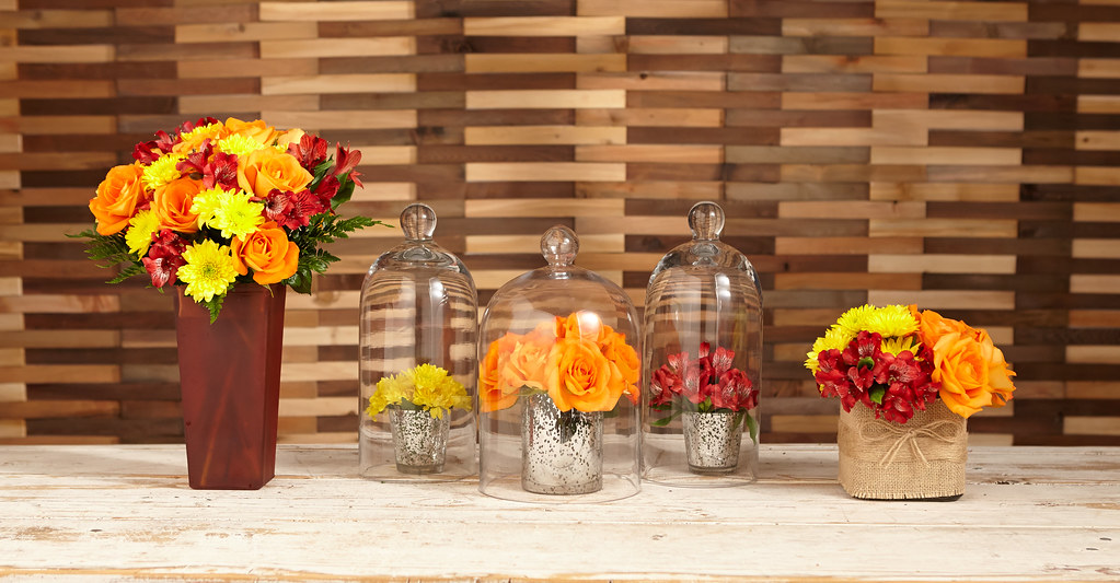 Three different ways to display orange roses red alstroemeria Peruvian lilies and yellow viking poms on a table including a tall orange vase, bell shaped glass and a canvas burlap short vase