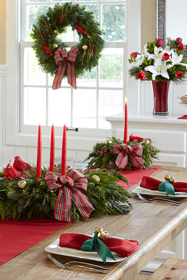 pine wreath hanging in the window behind a table set with red napkins and pine centerpieces with candles and ribbons and a boquet of red and white flowers in a glass vase on the mantel