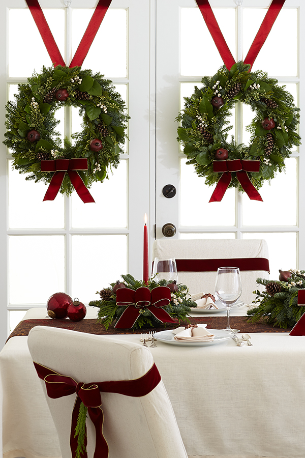two pine wreaths hanging over french door windows behind a table set with candle centerpieces and ribbons with a white tablecloth on the table