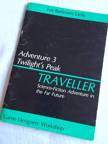 A photograph of the cover of Twilight’s Peak, an adventure for Traveller. A5 sized, the book is black with green stripes and top and bottom. The top stripe has the words ‘for referees only’ and the bottom strip has the publisher, ‘Game Designer’s Workshop’. across the middle of the book is the signature Traveller logo with a green line, and the words ‘Adventure 3 - Twilight’s Peak’. The book is worn from use and lies on a white fabric background.