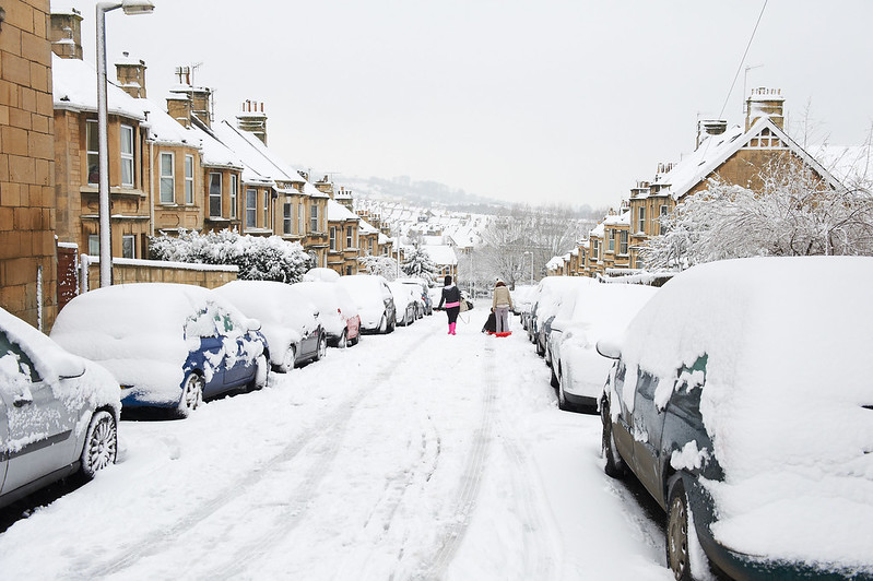 A snow-covered street lined with parked cars. A group of people pulls a sledge behind them in the distance.