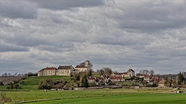 Hilltop Burgundy village with stone houses and a church tower, green fields in the foreground, and a cloudy sky.