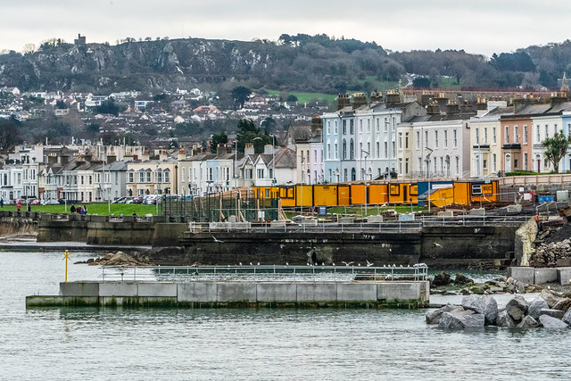 Harbourfront in Dún Laoghaire with pastel townhouses along the promenade, orange construction cabins, and a rocky breakwater along the calm water.