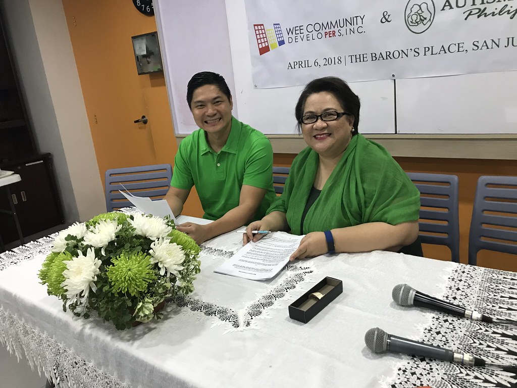 The image shows Ms. Mona wearing black outfit with green long scarf spreaded out and Ms. Cesar Wee wearing green collared shirt in front of the table during MOU signing.