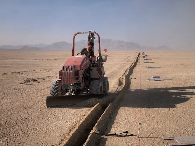 Red tracked tractor with a front blade plows a trench across cracked desert; a line of survey markers and equipment runs along the trench toward distant mountains.