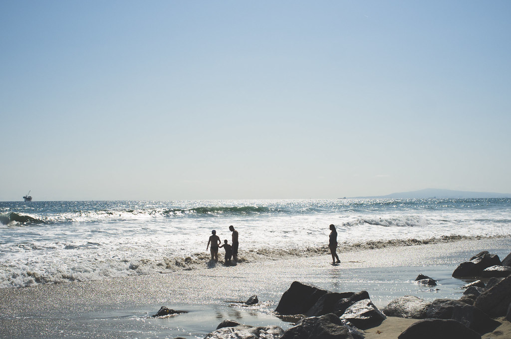Silhouetted family and beachgoers wading in sparkling ocean surf along a rocky California coastline.