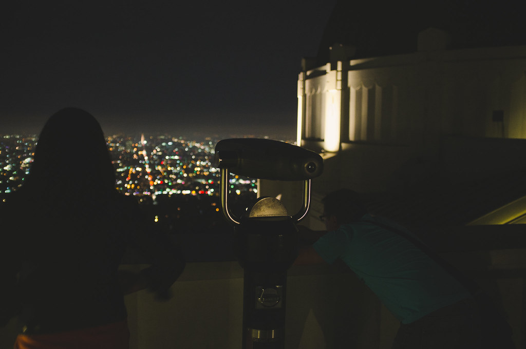 Visitors by an observation telescope on Griffith Observatory deck overlooking Los Angeles lights.