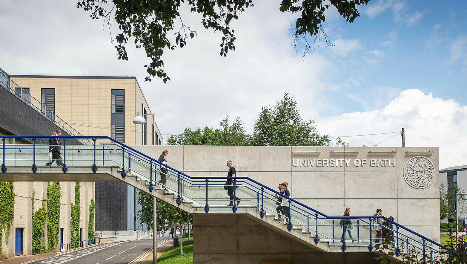 people walking over a bridge with University of Bath buildings and logo in the background