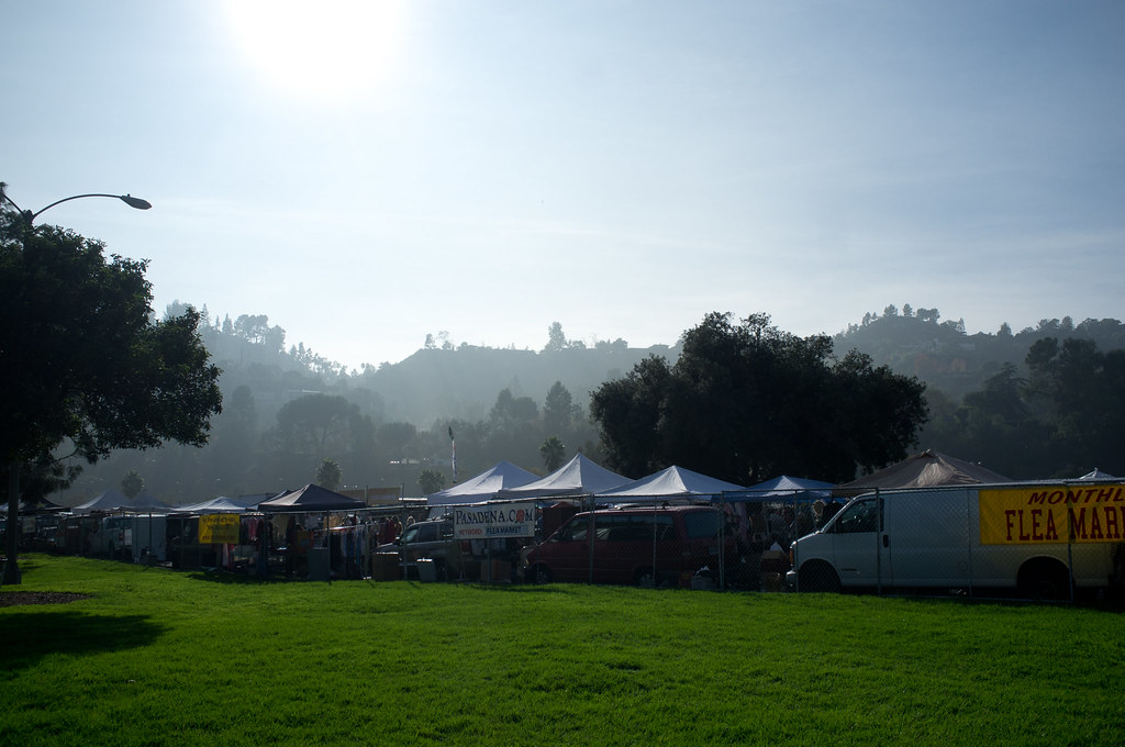 Open grass lawn overlooking white vendor canopies and vans at the Pasadena flea market beneath hazy morning hills.
