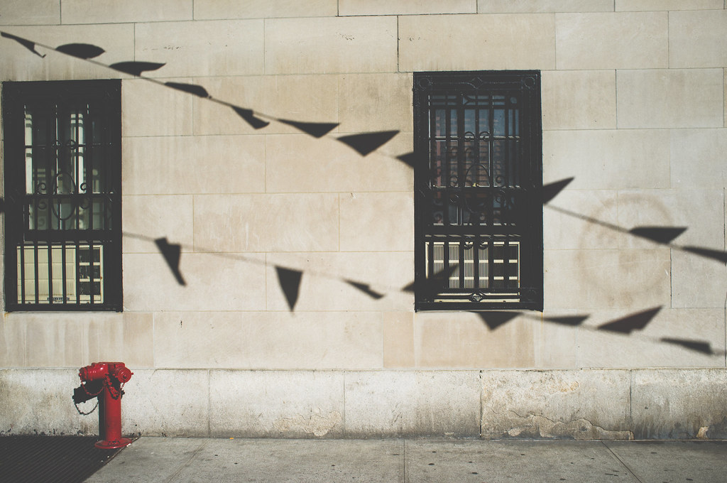 Pennant banners casting diagonal shadows on a stone facade with barred windows and a red fire hydrant.