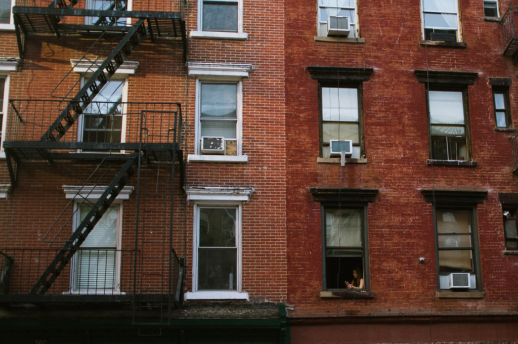 Red brick tenement buildings in NYC with black fire escapes and a resident looking out a lower window.