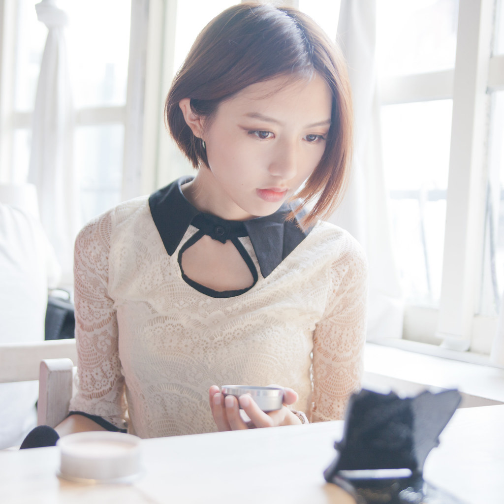 一位女子坐在窗邊的白色桌前，低頭看著手上的小圓盒，身後是白色窗框，沒有看鏡頭