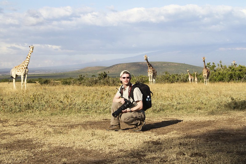 A white male wearing a cap and holding a camera with a long lens, kneeling in front of a number of giraffes