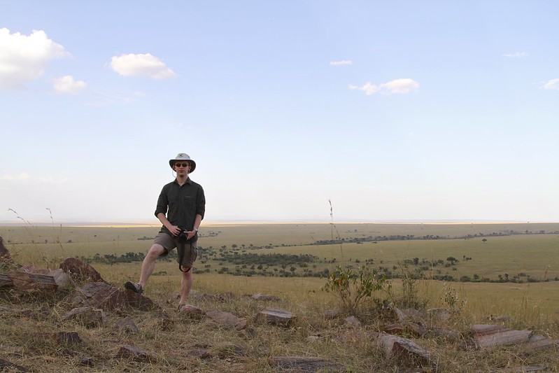 White male wearing safari clothes and hat, holding a camera, with the expansive Masai Mara all around
