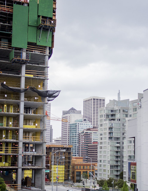 Under-construction high-rise with green panels on left and modern glass towers on the right under a cloudy sky.