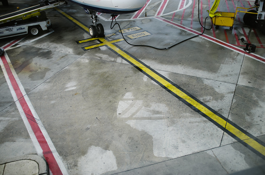 Airport window view of painted yellow lines and nose landing gear of a JetBlue airliner on the tarmac.