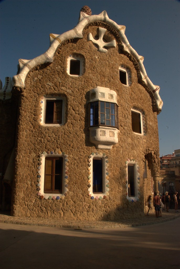 A photo of Parc Guell, which is a large medium-brown structure with a while roof and carved out rectangular windows. 