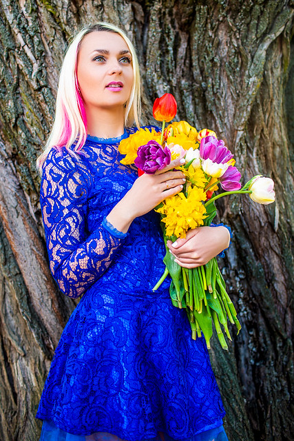 Fashion Concepts and Ideas. Positive Caucasian Girl with Bunch of Fresh Colorful Tulips Posing Against Old Brown Tree Behind. Model in Blue Dress During Spring Time.