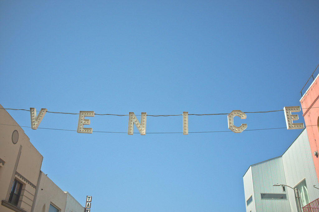 Illuminated 'VENICE' sign hanging from overhead cables across the street against a vivid blue sky.