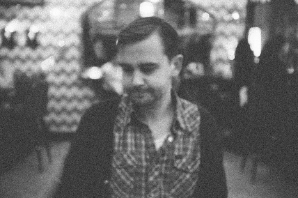 Portrait of a man in a plaid shirt looking downward against chevron patterned wallpaper.
