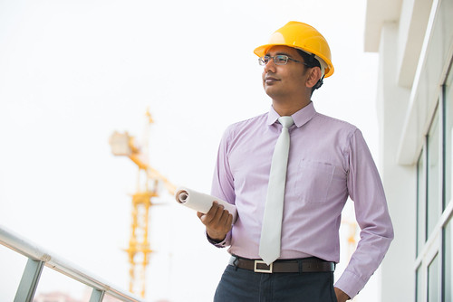 indian male architect holding plans with construction background