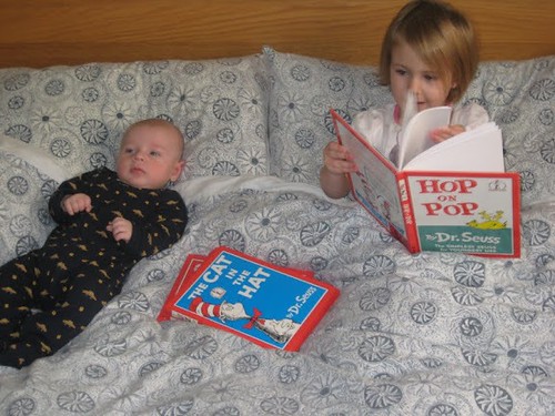 A young girl sitting in bed with several Dr. Seuss books and reading to her infant brother.