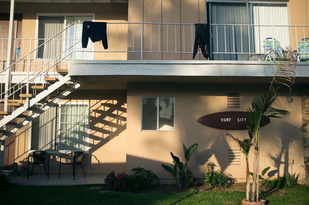 Sunny apartment building with outdoor staircase shadows and a wooden 'SURF CITY' surfboard plaque.