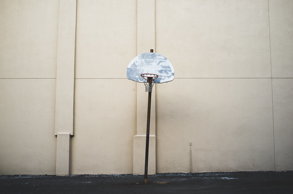 Weathered basketball hoop and backboard mounted on a tall pole against a plain beige building exterior wall.
