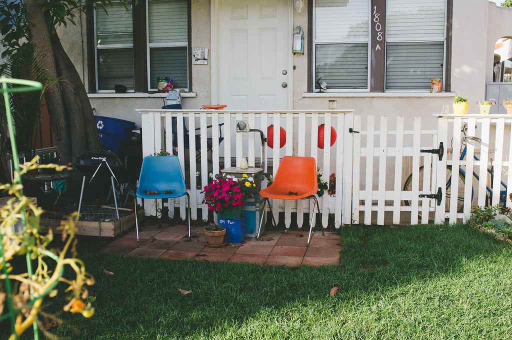 Sunny patio with blue and orange fiberglass shell chairs, blooming pink flowers, and a grill.