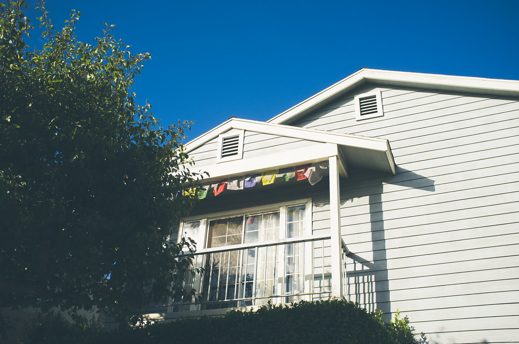 Upper balcony of a white house decorated with colorful Tibetan prayer flags under a blue sky.