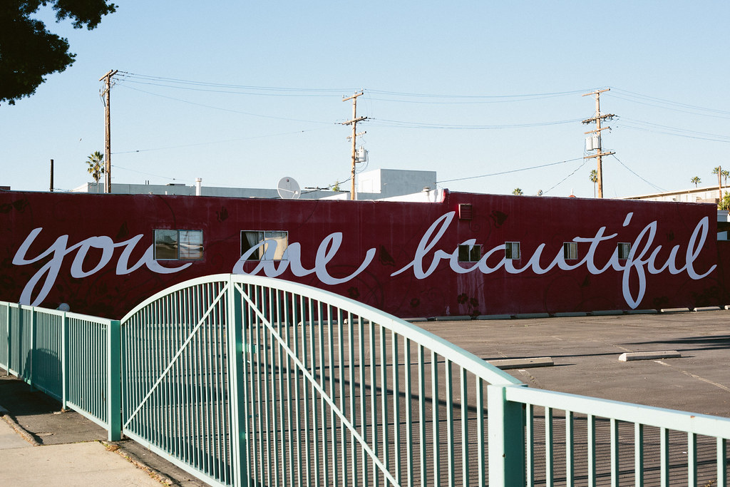 Deep red building behind a teal gate painted with white cursive script reading 'you are beautiful'.