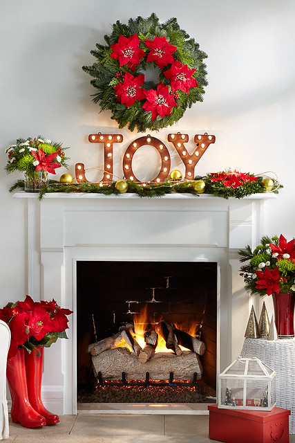 Merry mantel with lighted Joy sign pine cuttings with ornaments a centerpiece boots vase with poinsettias red glass vase with mums and pine wreath over a fireplace and a Christmas terrarium on a red box