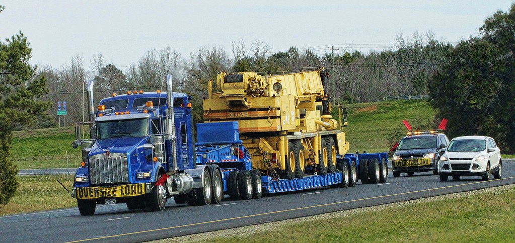 Kenworth trailer truck hauling an excavator on a highway. The truck has yellow signs that read oversize load.