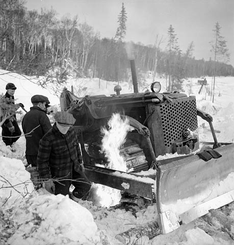 Lumbermen gather around a fire built under a frozen snowplow to thaw it out, Sloe Lake, Quebec / Des bûcherons se rassemblent autour d’un feu allumé sous un chasse-neige pour le faire dégeler, lac Sloe (Québec)